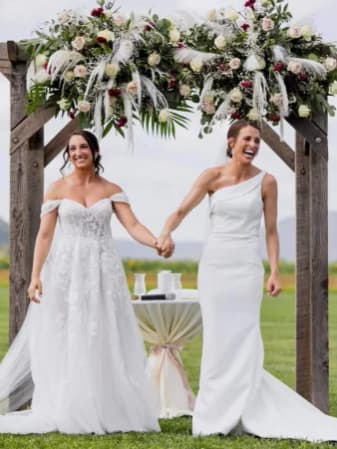 Two brides holding hands beneath a wooden arch dressed with white roses and greenery
